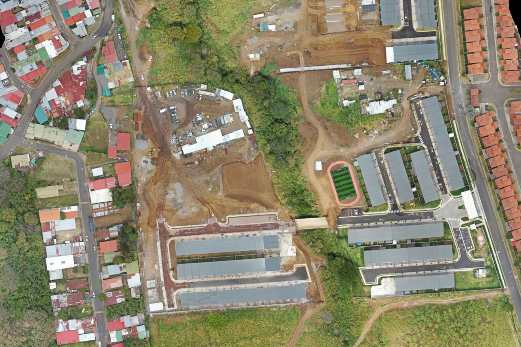 Ortofoto vertical del Condominio UNIKO en construcción: bloques de vivienda con techos grises alineados junto a una calle interna, filas de casas de teja al otro lado de la vía y movimiento de tierras en la parcela contigua.